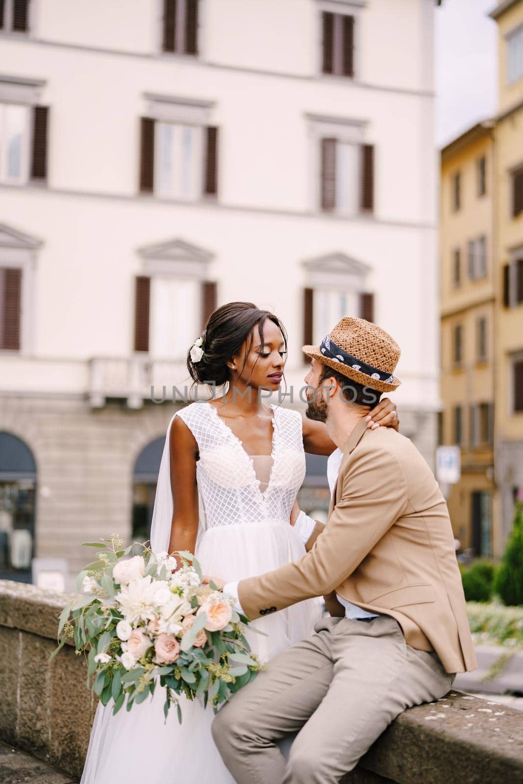 African-American bride in a white dress with a long veil and bouquet, and Caucasian groom in a sand jacket and straw hat. Interracial wedding couple. Wedding in Florence, Italy.