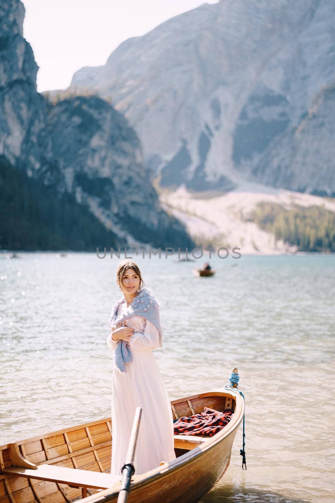 A bride stands in a wooden boat at the Lago di Braies in Italy. Wedding in Europe, on Braies lake. A young girl in a white wedding dress, covers her shoulders with a blue sweater. by Nadtochiy