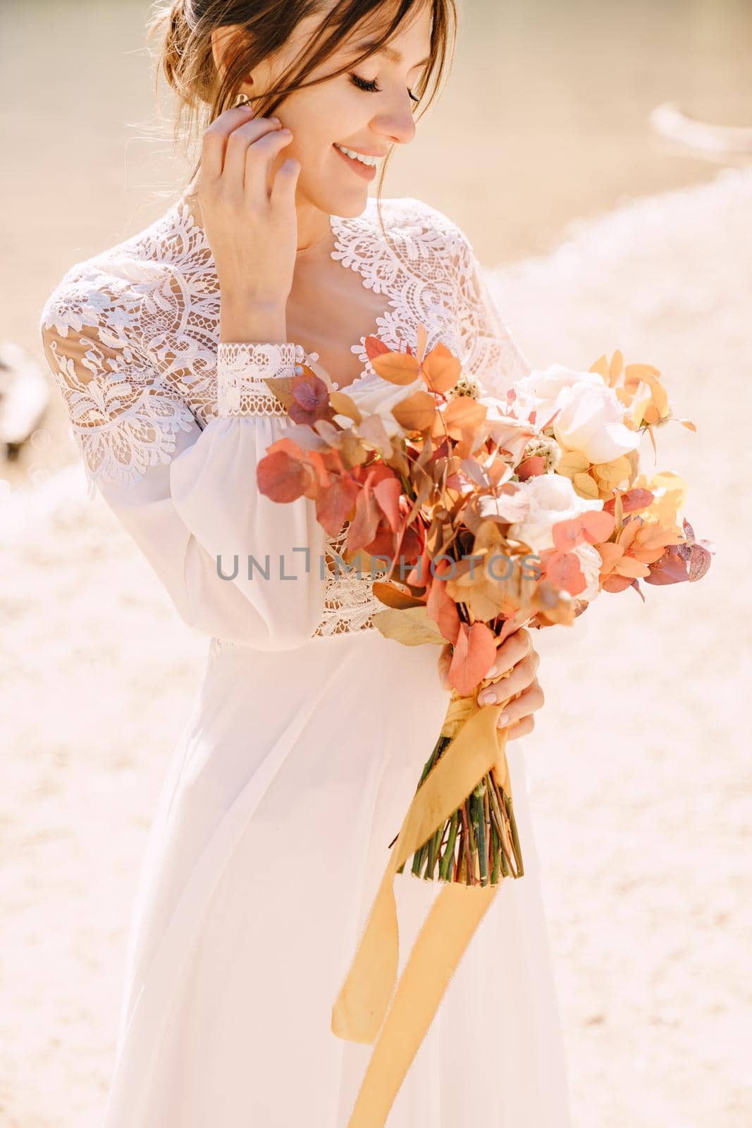 Beautiful bride in a white dress with sleeves and lace, with a yellow autumn bouquet of dried flowers and peony roses, on the Lago di Braies in Italy. Destination wedding in Europe, on Braies lake. by Nadtochiy