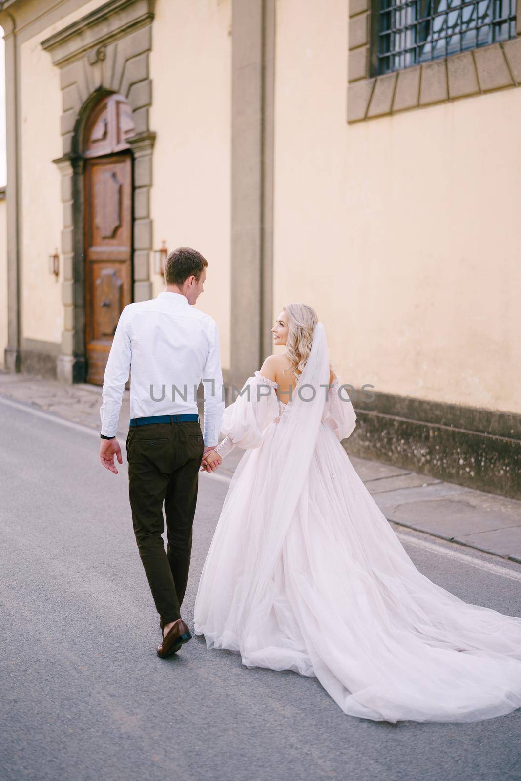 Beautiful bride and groom walking hand in hand away from the camera outside of the old villa in Italy, in Tuscany, near Florence.
