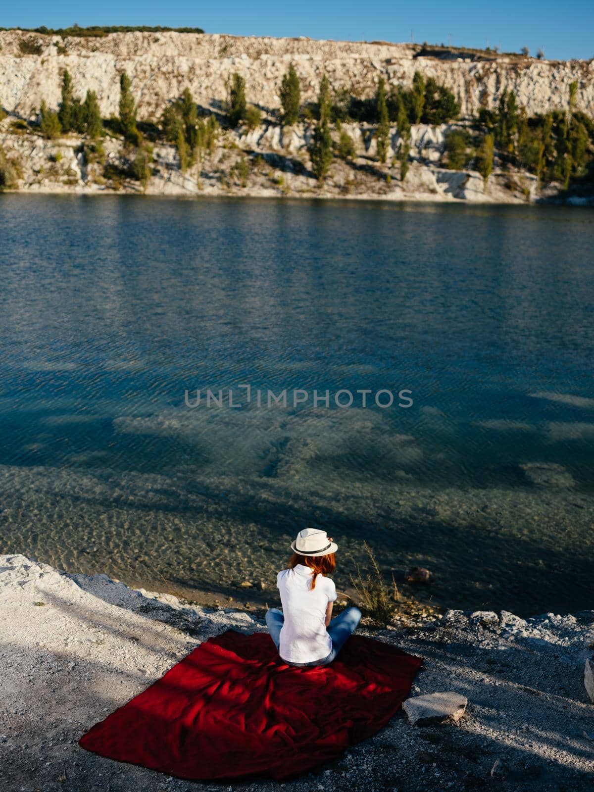 A woman in a white T-shirt sits on a red cloth near the sea in nature and a hat on her head by SHOTPRIME