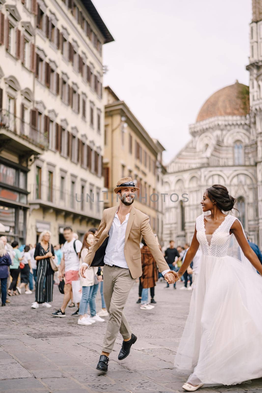 Interracial wedding couple. Wedding in Florence, Italy. African-American bride and Caucasian groom run along Piazza del Duomo. by Nadtochiy