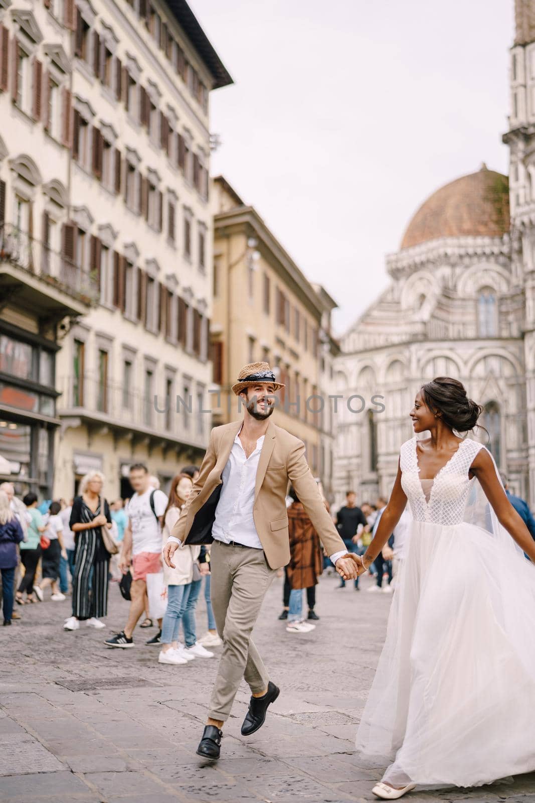 African-American bride and Caucasian groom run along Piazza del Duomo. Multiracial wedding couple. Wedding in Florence, Italy.