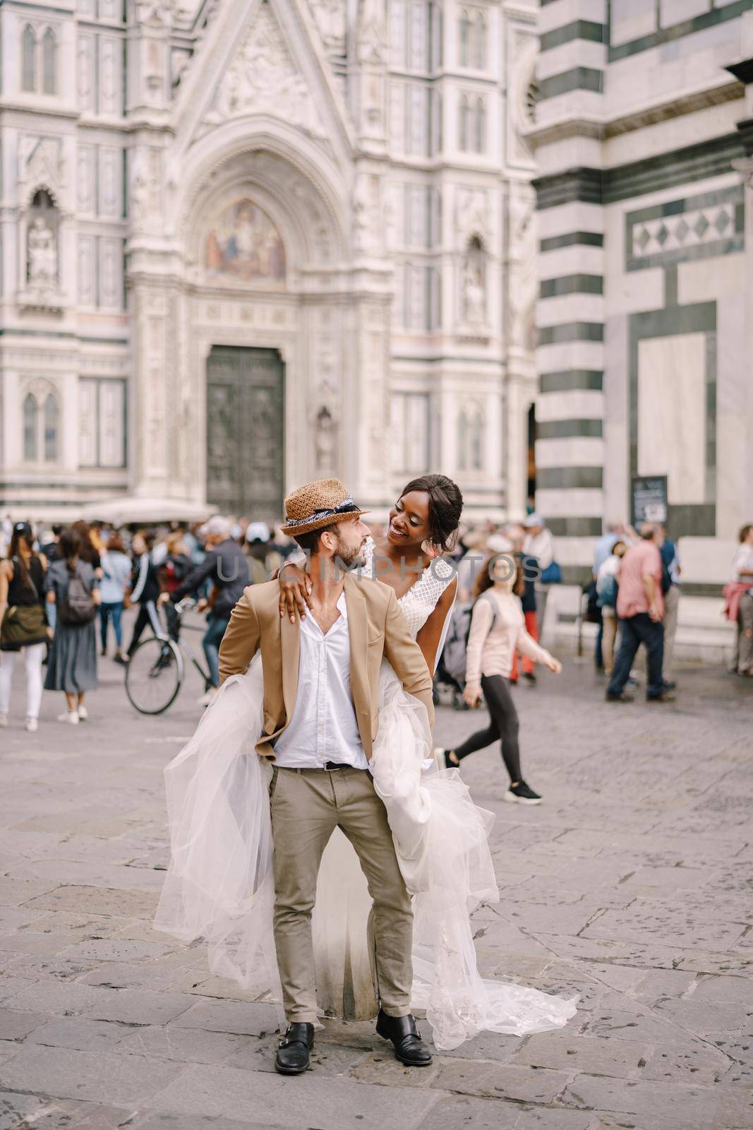 Multiethnic wedding couple. Wedding in Florence, Italy. An African-American bride hopped onto the shoulders of a Caucasian groom in the Piazza del Duomo. by Nadtochiy