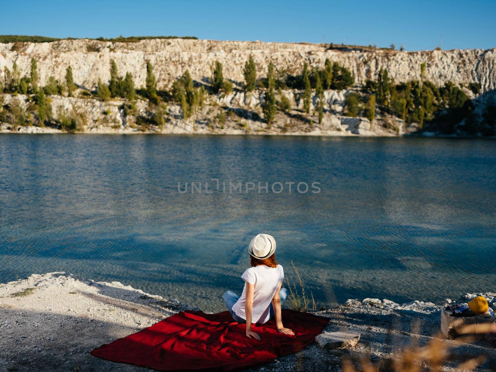 A traveler wearing a hat sits on a red cloth near the sea in nature by SHOTPRIME