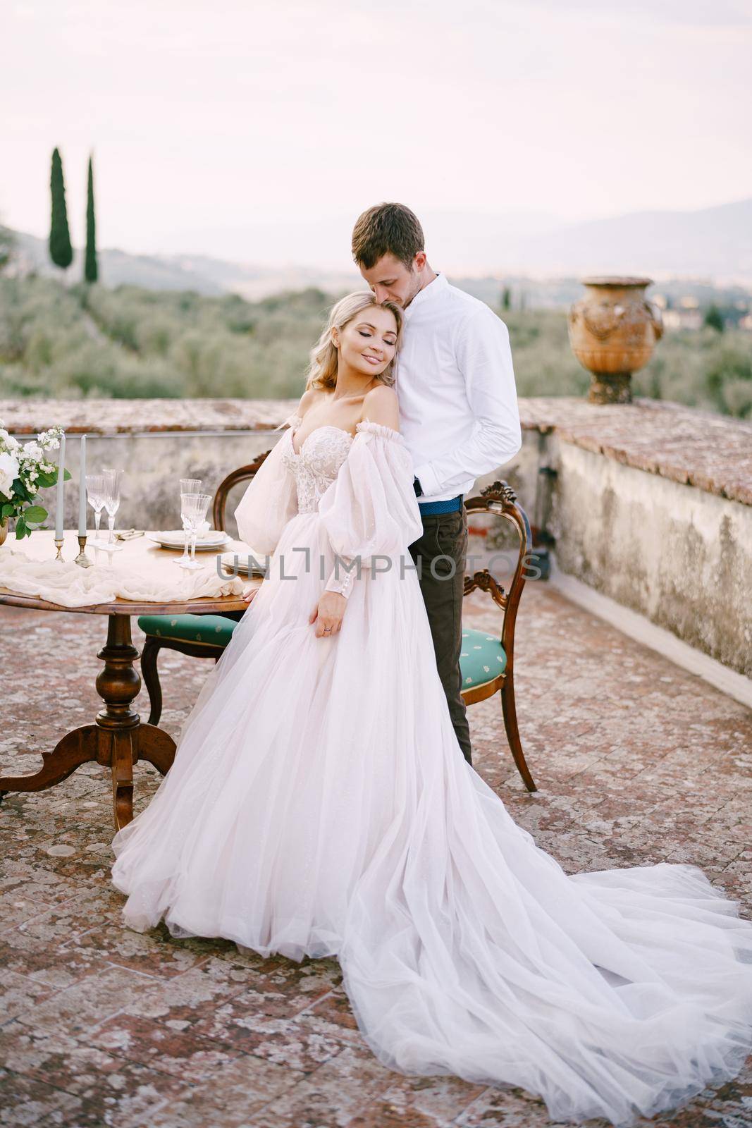 Wedding at an old winery villa in Tuscany, Italy. A wedding couple is standing near the table for a wedding dinner, the groom hugs the bride by the waist. by Nadtochiy