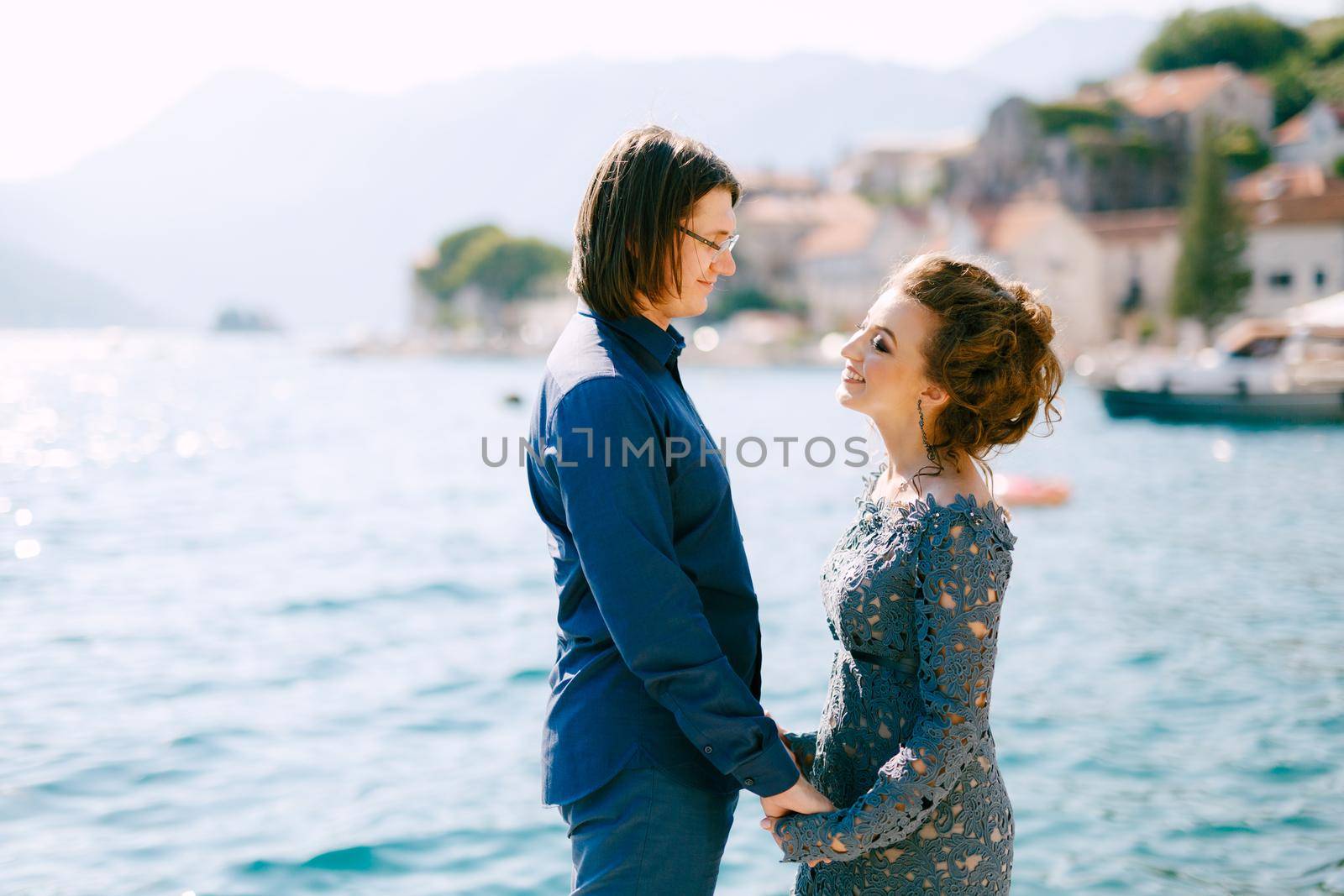 The bride in a stylish grey wedding dress and the groom stand holding hands on the seashore near the old town of Perast, close-up. High quality photo