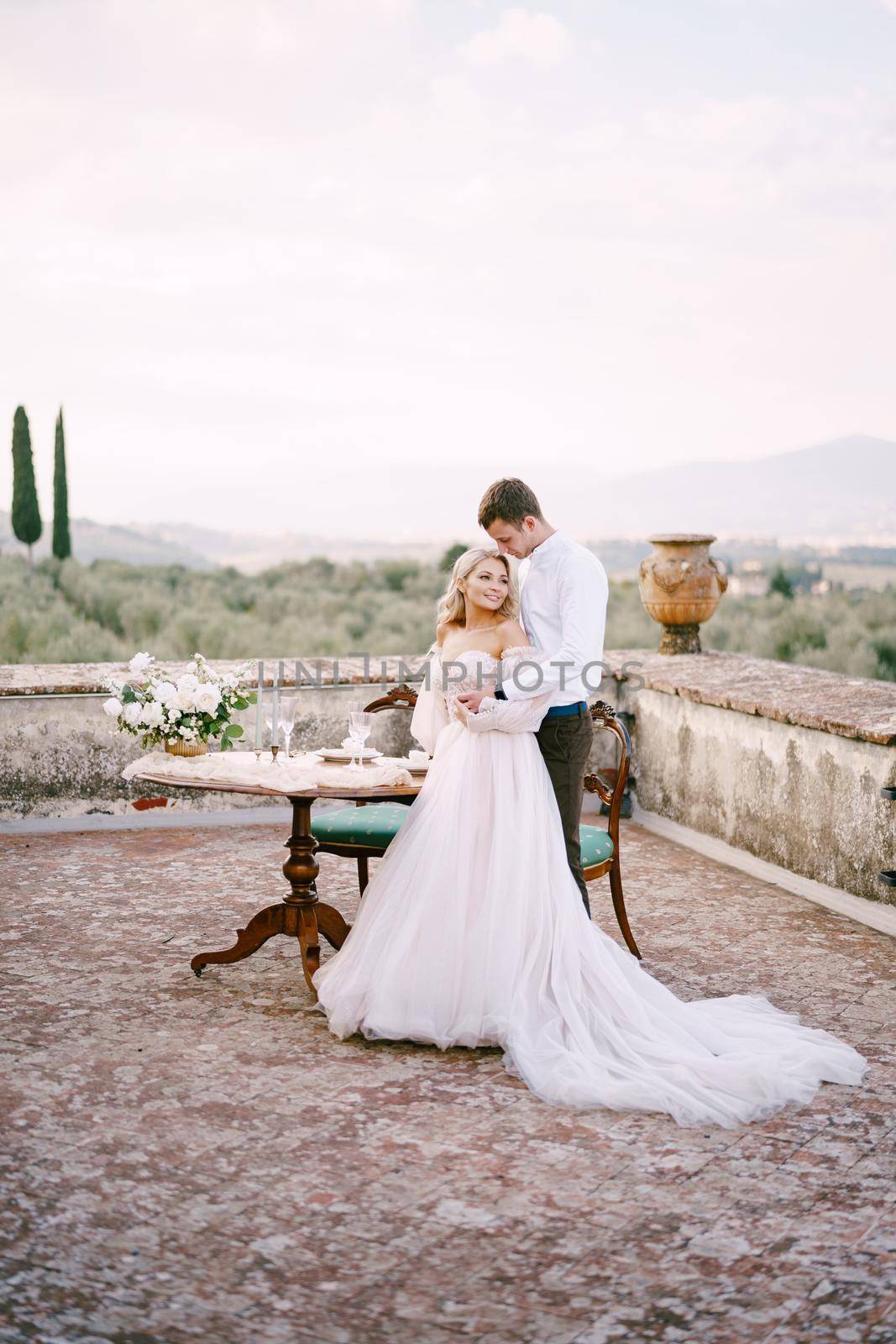 Wedding at an old winery villa in Tuscany, Italy. The wedding couple stands near the table for the wedding dinner, the groom hugs the bride at the waist.