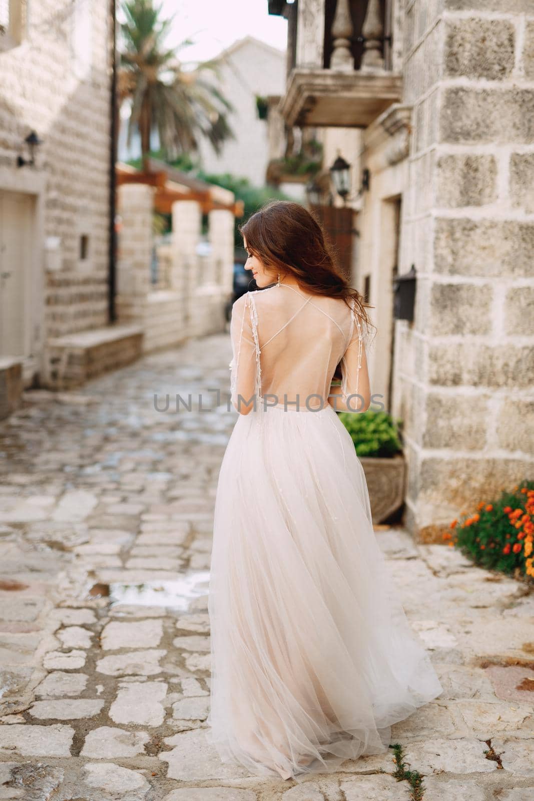 A bride in a stylish dress with an open back stands in front of an ancient building on a cozy Perast street, back view by Nadtochiy