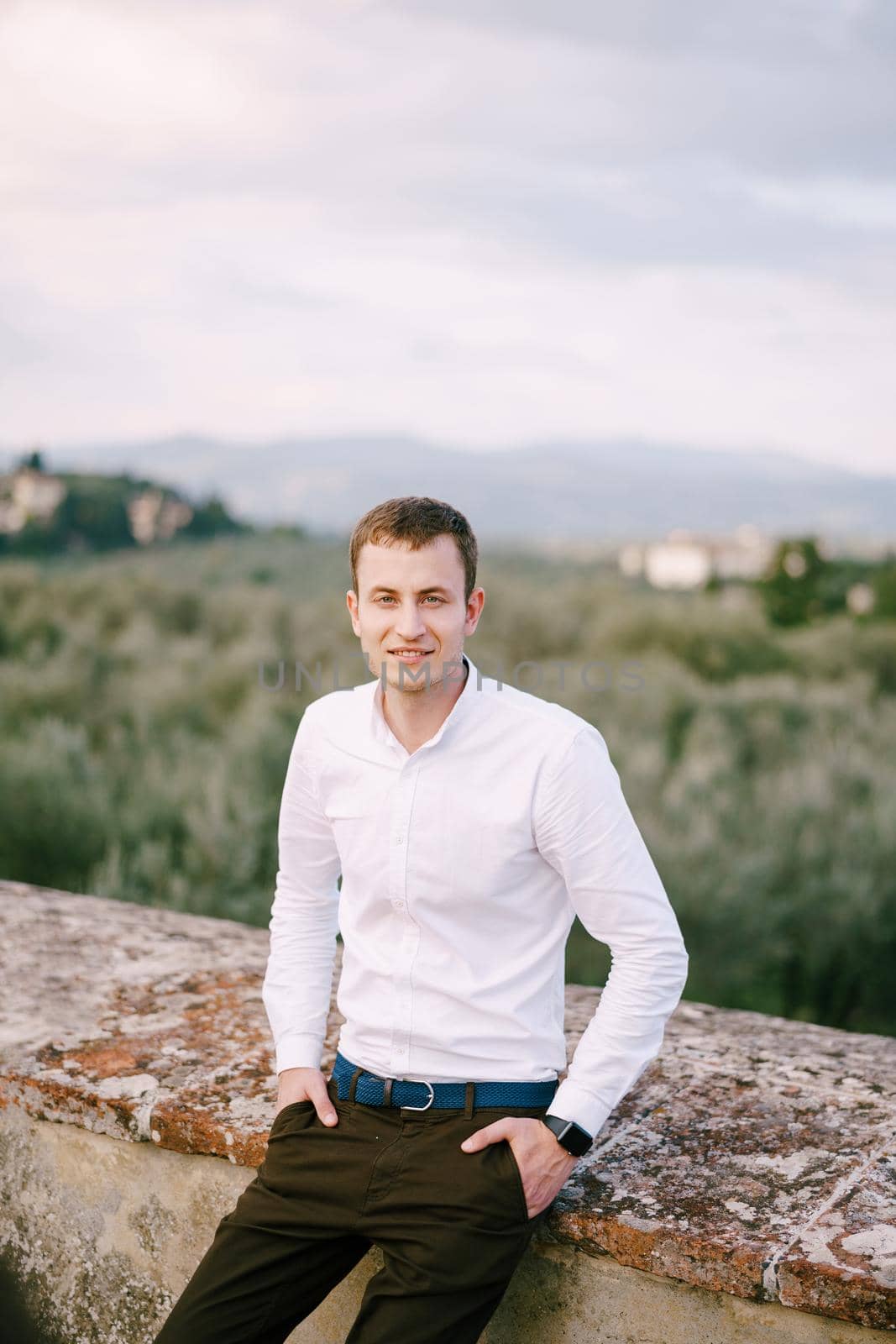 A large male portrait on the roof of an old winery villa in Tuscany, Italy. White long-sleeved shirt, smart watch, blue belt, hands in pants pockets.