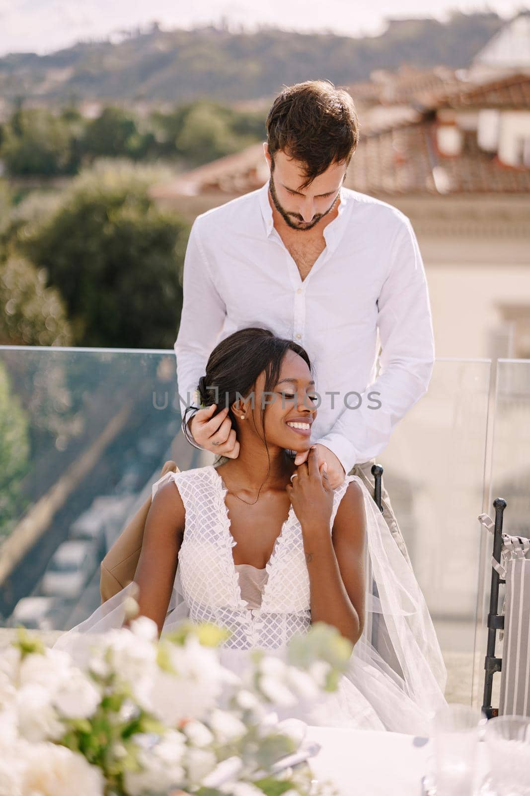 Interracial wedding couple. Destination fine-art wedding in Florence, Italy. African-American bride sits at wedding table, Caucasian groom hugs her shoulders.