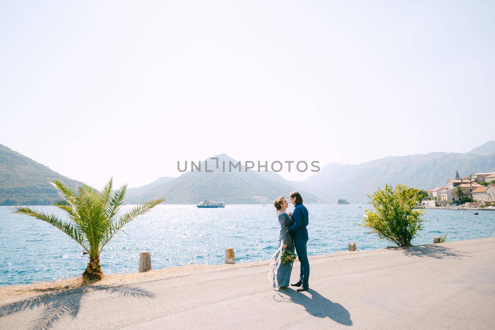 The bride in stylish grey dress and groom hugging on the pier near the old town of Perast . High quality photo