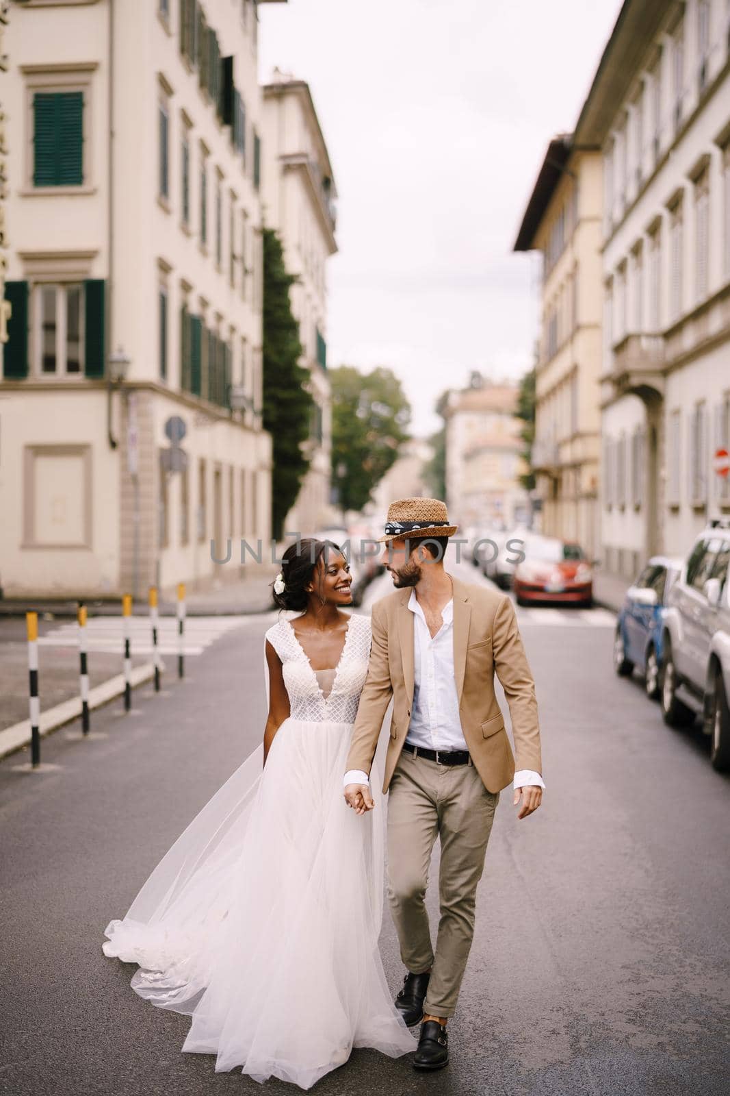 Wedding in Florence, Italy. Multiethnic wedding couple. African-American bride in a white dress and Caucasian groom in a hat are walking along the road among cars. by Nadtochiy