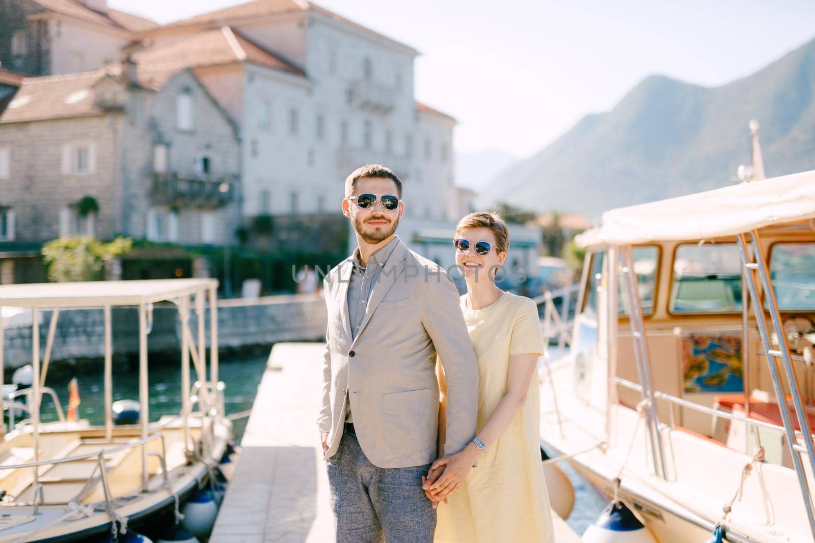 A man and a woman in sunglasses stand hugging each other on the pier near the tourist boats and hold hands . High quality photo