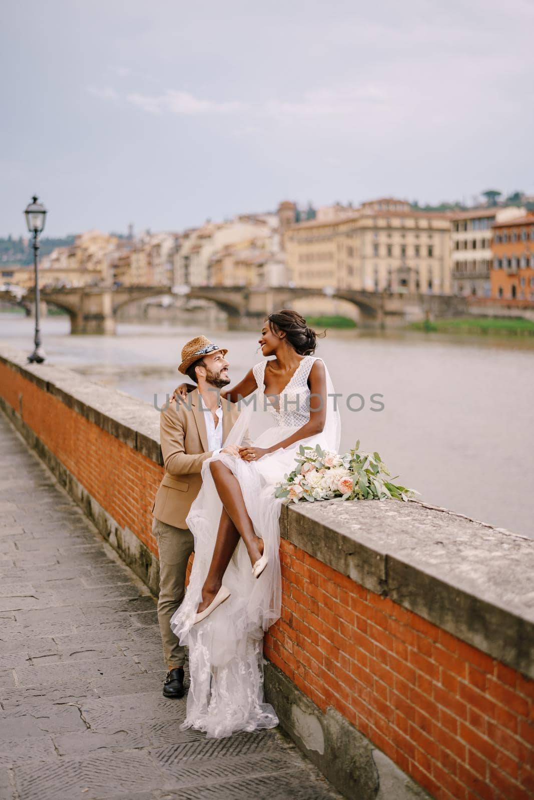 An African-American bride sits on a brick wall and a Caucasian groom hugs her. The embankment of the Arno River, overlooking city and bridges. Interracial wedding couple. Wedding in Florence, Italy.