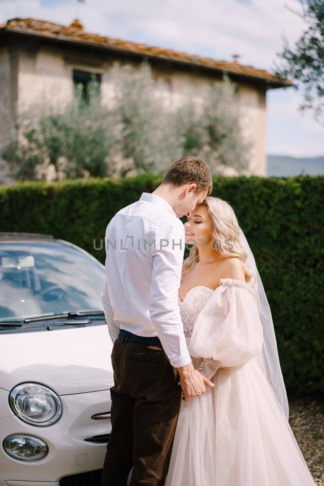 Beautiful bride and groom looking at each other and holding hands in front of a convertible at the Villa in Tuscany, Italy by Nadtochiy