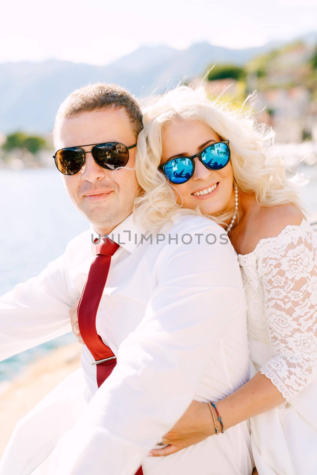 The bride and groom in sunglasses sit embracing on the pier in Perast, the bride is snuggling against the back of the groom . High quality photo