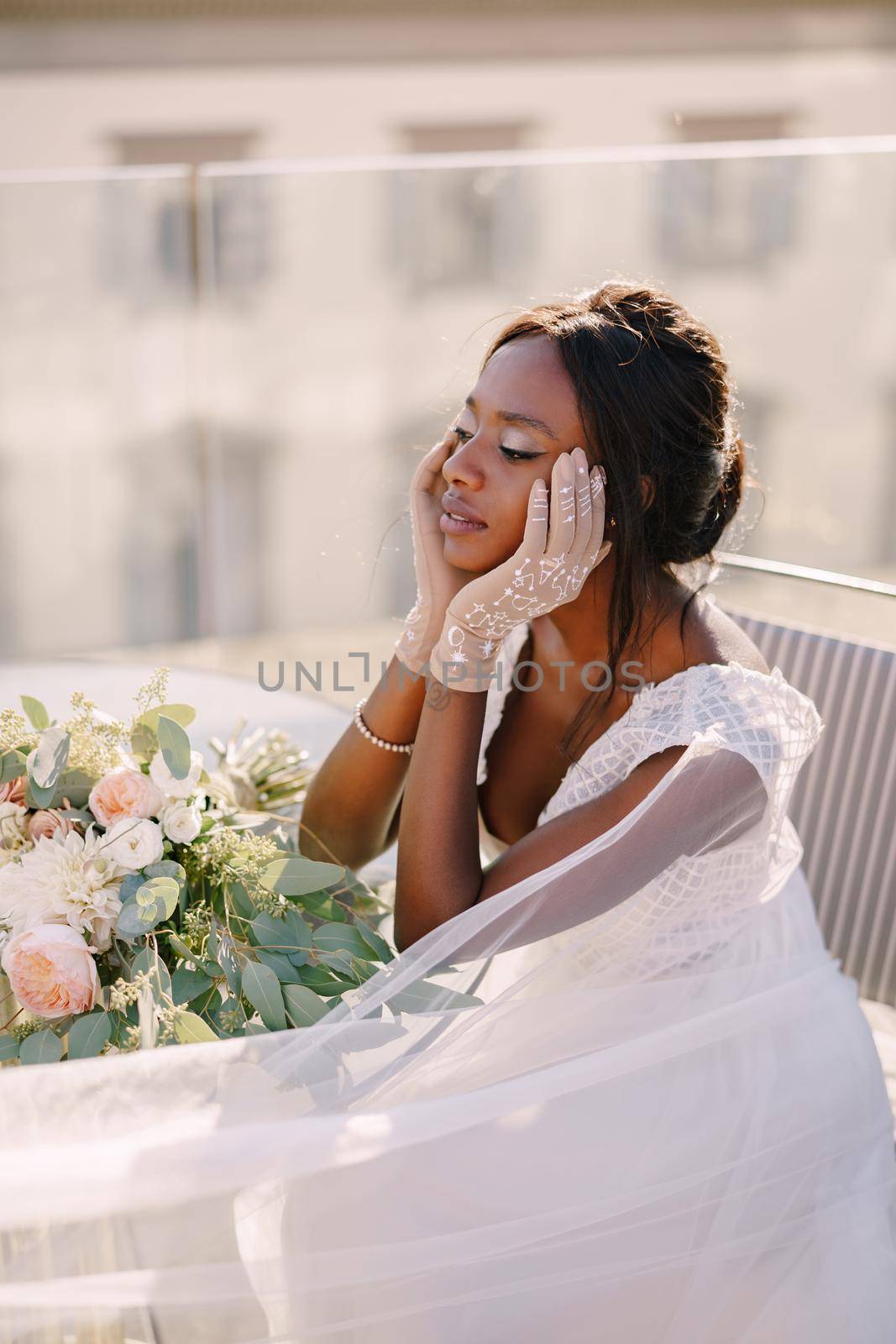 Destination fine-art wedding in Florence, Italy. African-American bride sits at the table, touches his face with her hands in gloves, a bouquet lies on the table.