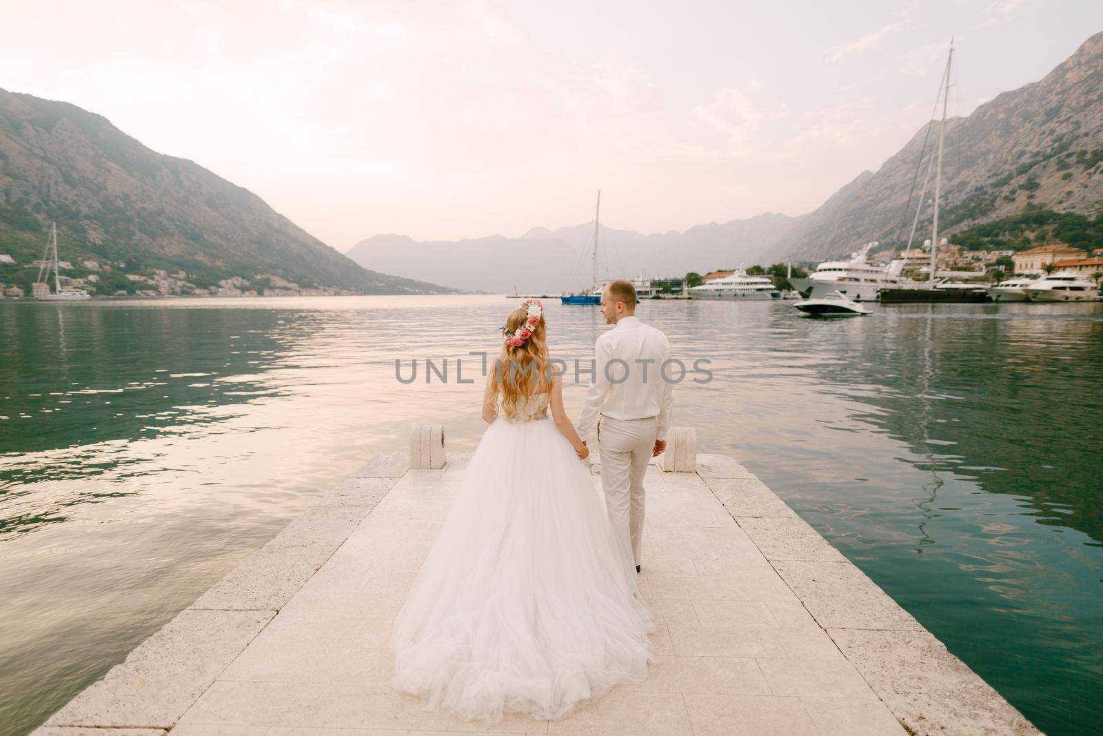 The bride in a wreath and groom walk along the pier holding hands near the old town of Kotor in the Bay of Kotor by Nadtochiy