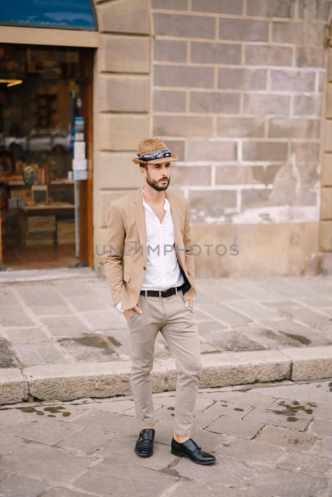 A young guy with a straw hat, a white unbuttoned shirt and a sand jacket, a small beard walks around the city of Florence. by Nadtochiy