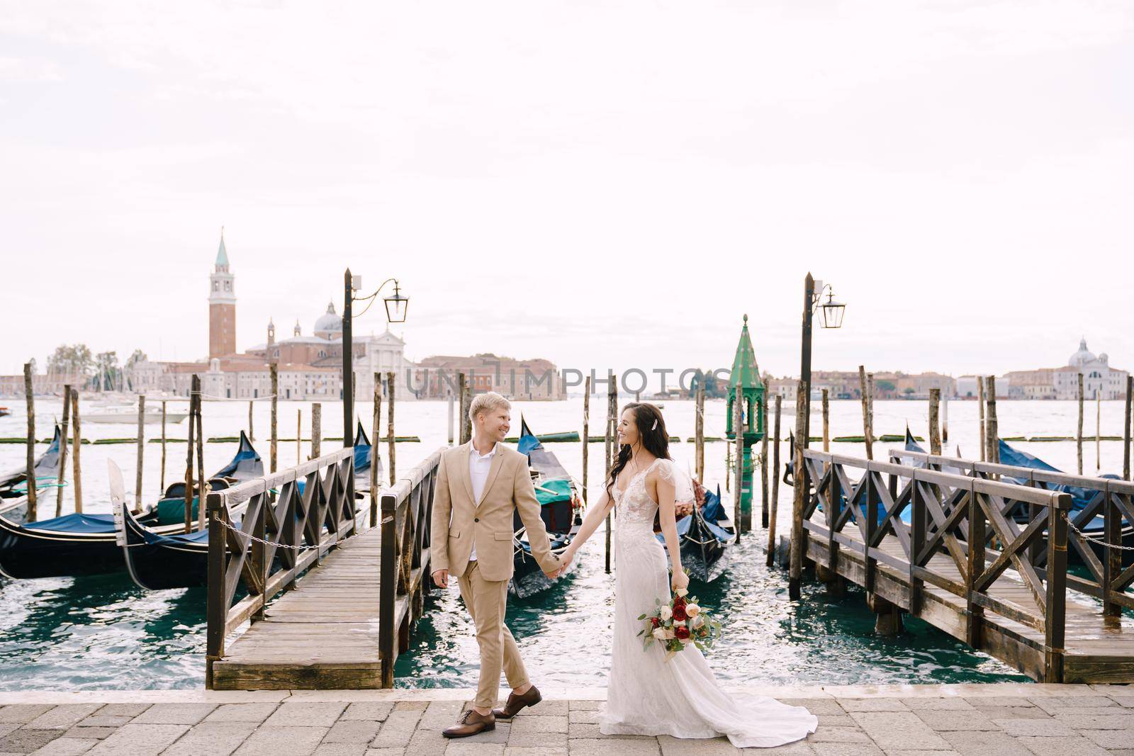 The bride and groom are walking along the gondola pier, holding hand in Venice, near Piazza San Marco, overlooking San Giorgio Maggiore and the sunset sky. The largest gondola pier in Venice, Italy. by Nadtochiy