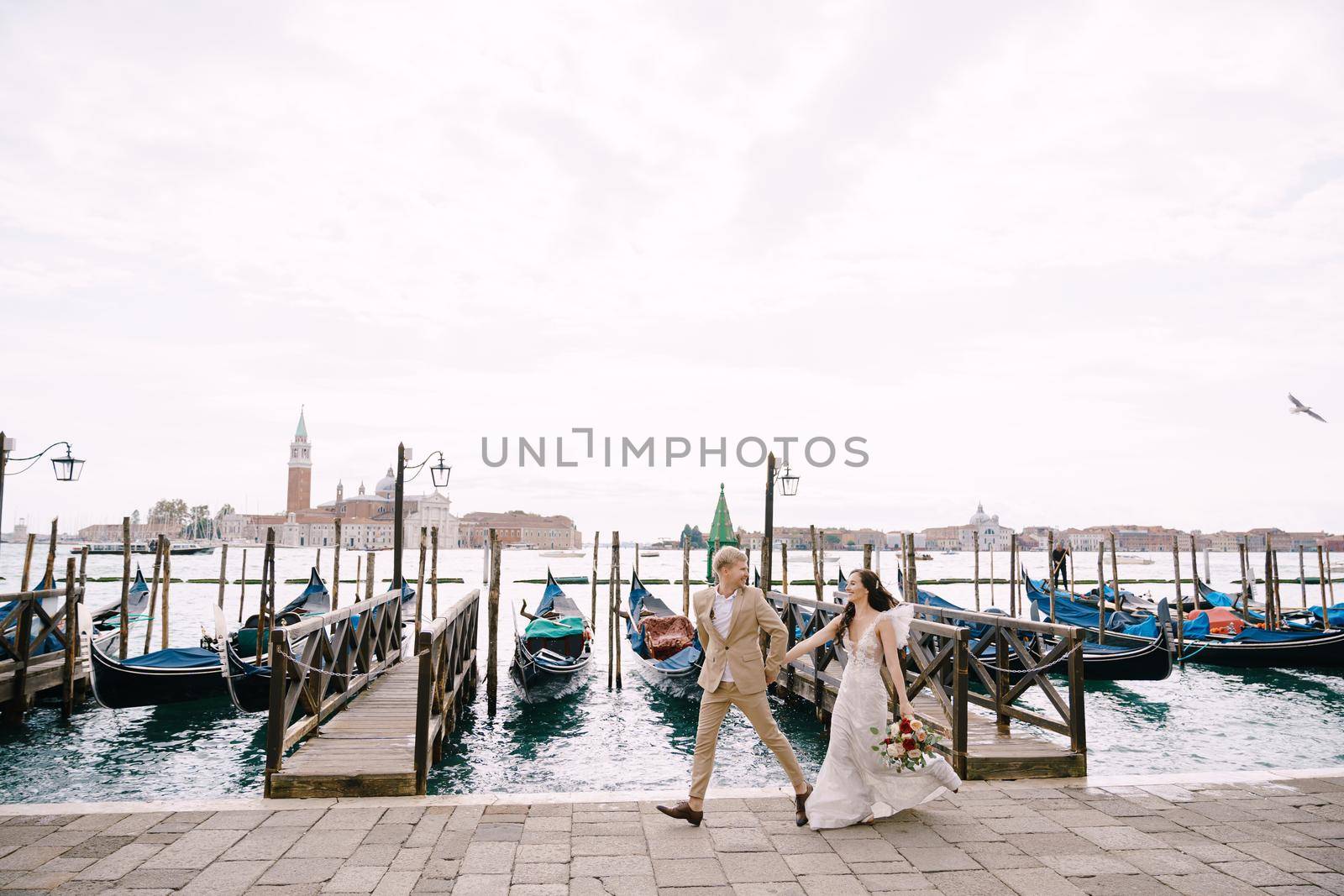 The bride and groom are run along the gondola pier, holding hand in Venice, near Piazza San Marco, overlooking San Giorgio Maggiore and the sunset sky. The largest gondola pier in Venice, Italy. by Nadtochiy