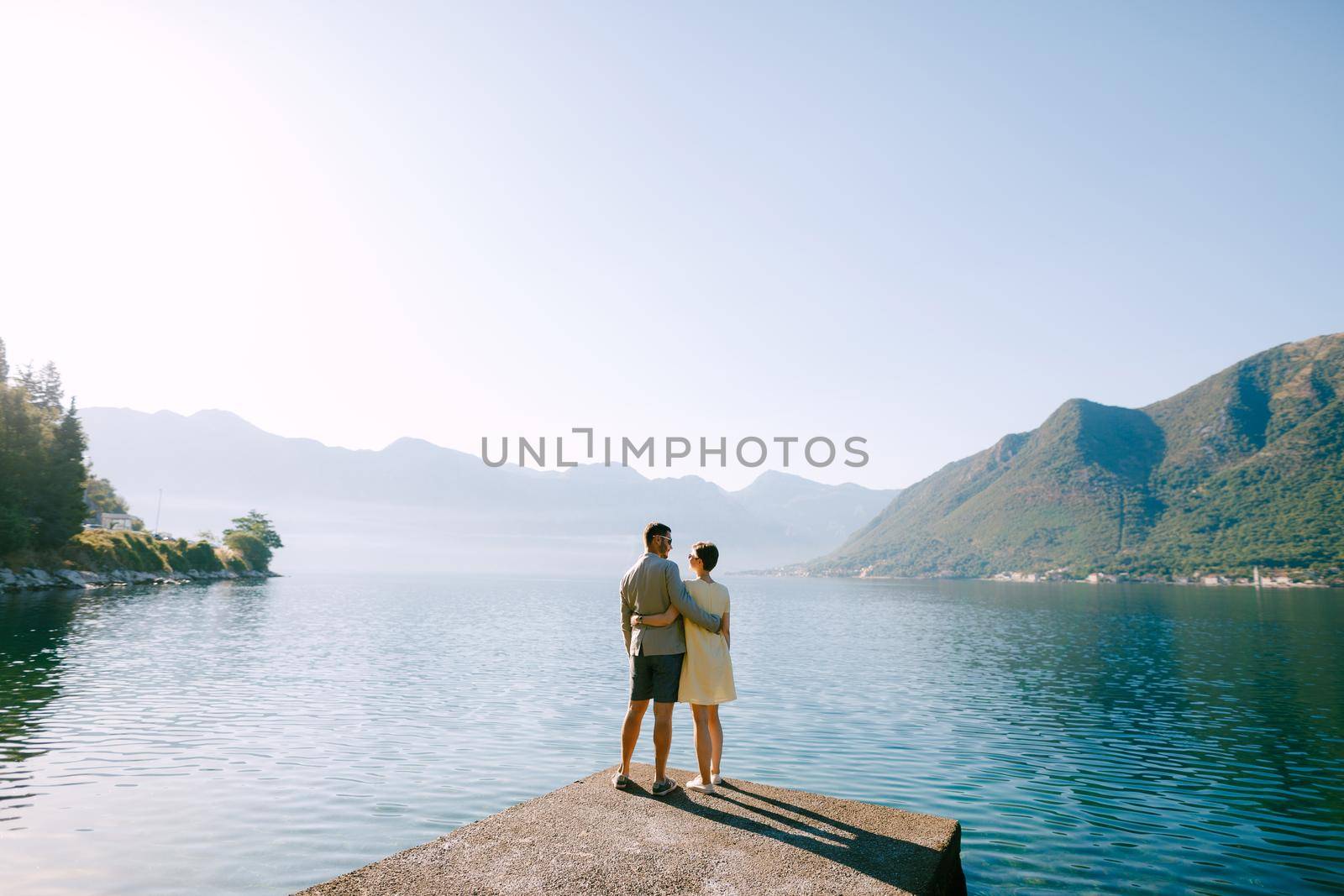 A couple in love stands hugging on a pier in the Bay of Kotor near Perast against the backdrop of mountains, back view by Nadtochiy