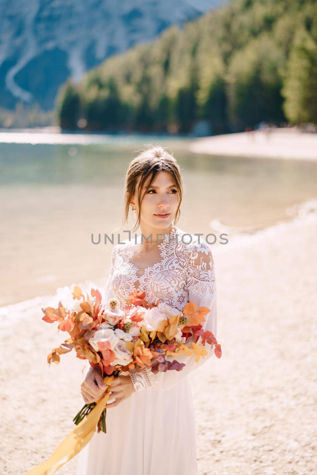 Beautiful bride in a white dress with sleeves and lace, with a yellow autumn bouquet of dried flowers and peony roses, at Lago di Braies in Italy. Destination wedding in Europe, at Braies lake.