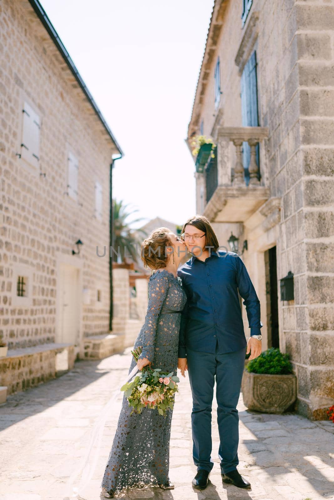 Bride in stylish blue dress kisses the groom against the background of beautiful white houses in the old town of Perast by Nadtochiy