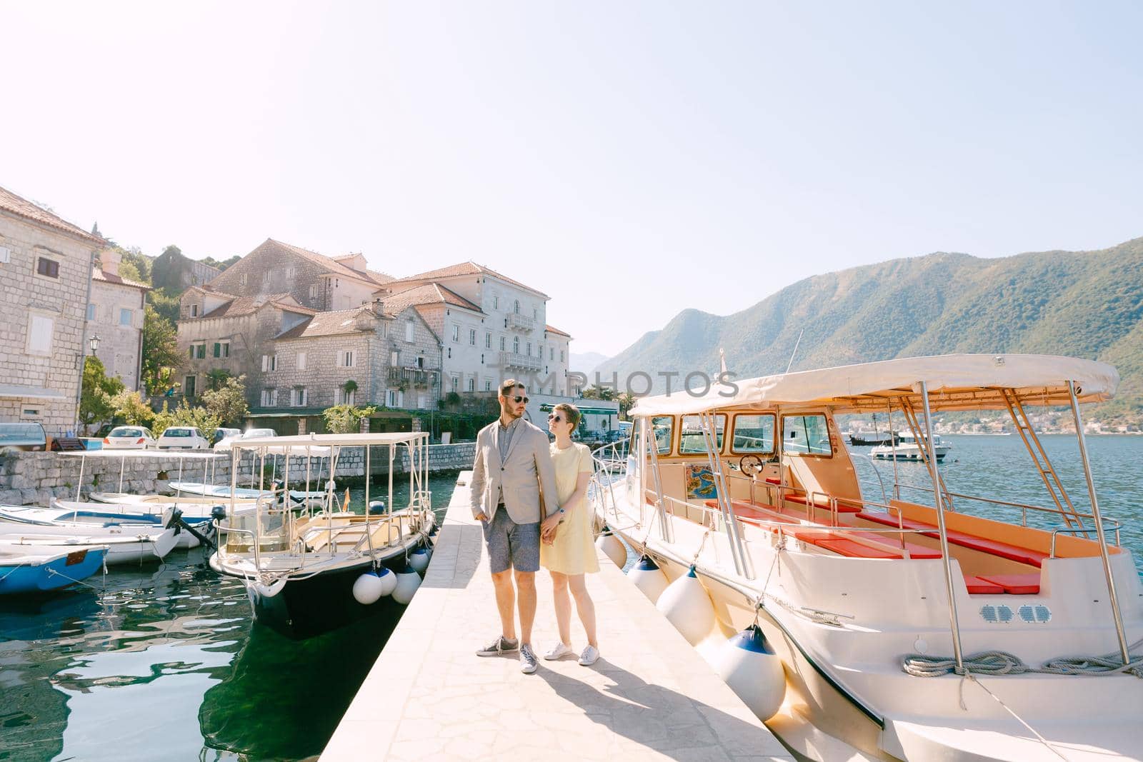 A man and a woman in sunglasses stand hugging each other on the pier near the tourist boats and hold hands . High quality photo