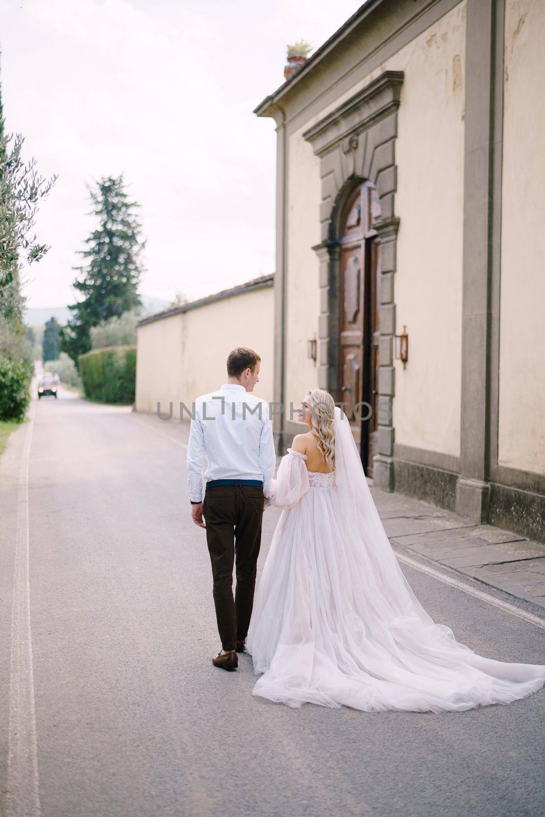 Beautiful bride and groom walking hand in hand away from the camera outside of the Villa in Tuscany, Italy by Nadtochiy