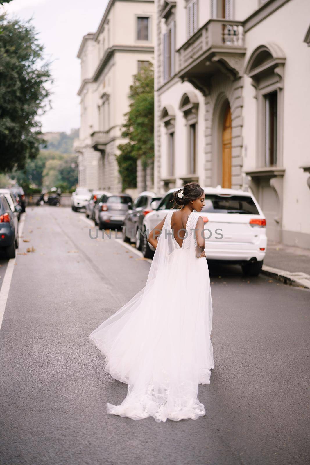 An African-American bride in a white dress, with a long veil, walks on the road on a city street. Wedding in Florence, Italy