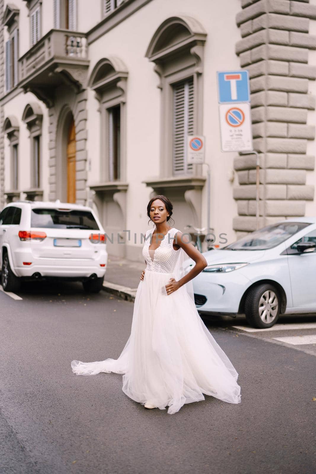 An African-American bride in a white dress, with a long veil, walks on the road on a city street. Wedding in Florence, Italy