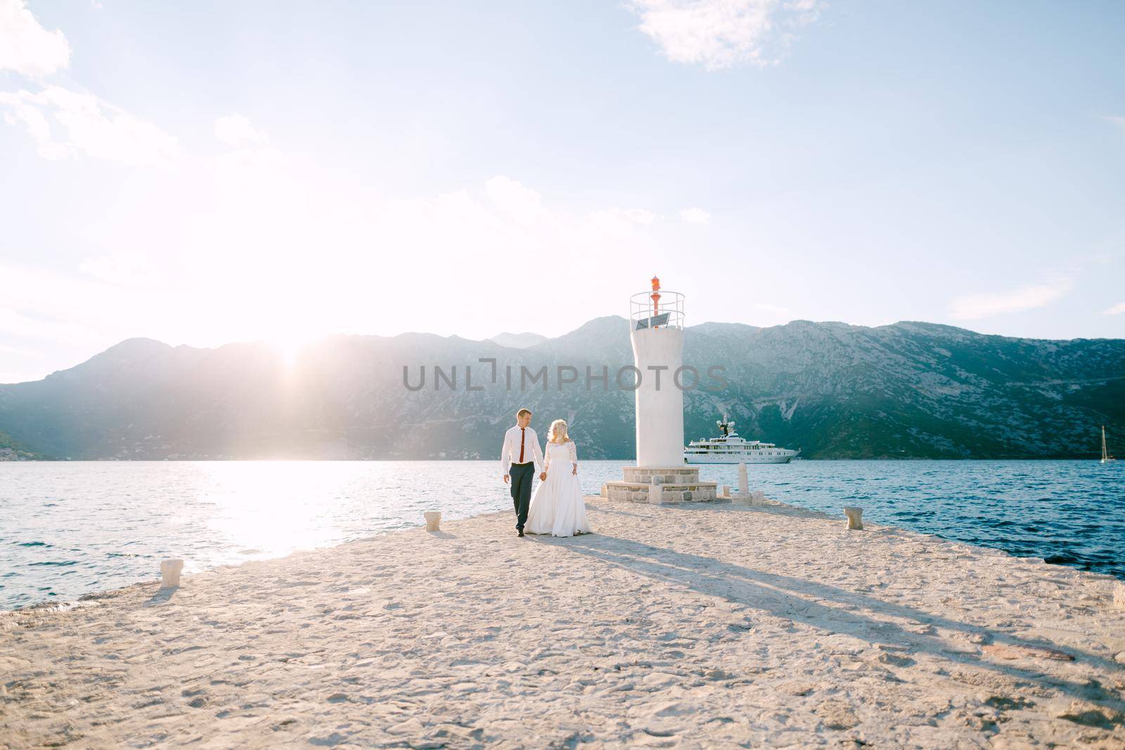 The bride and groom walk hand in hand near the lighthouse on the pier on the Our Lady of the Rocks island . High quality photo
