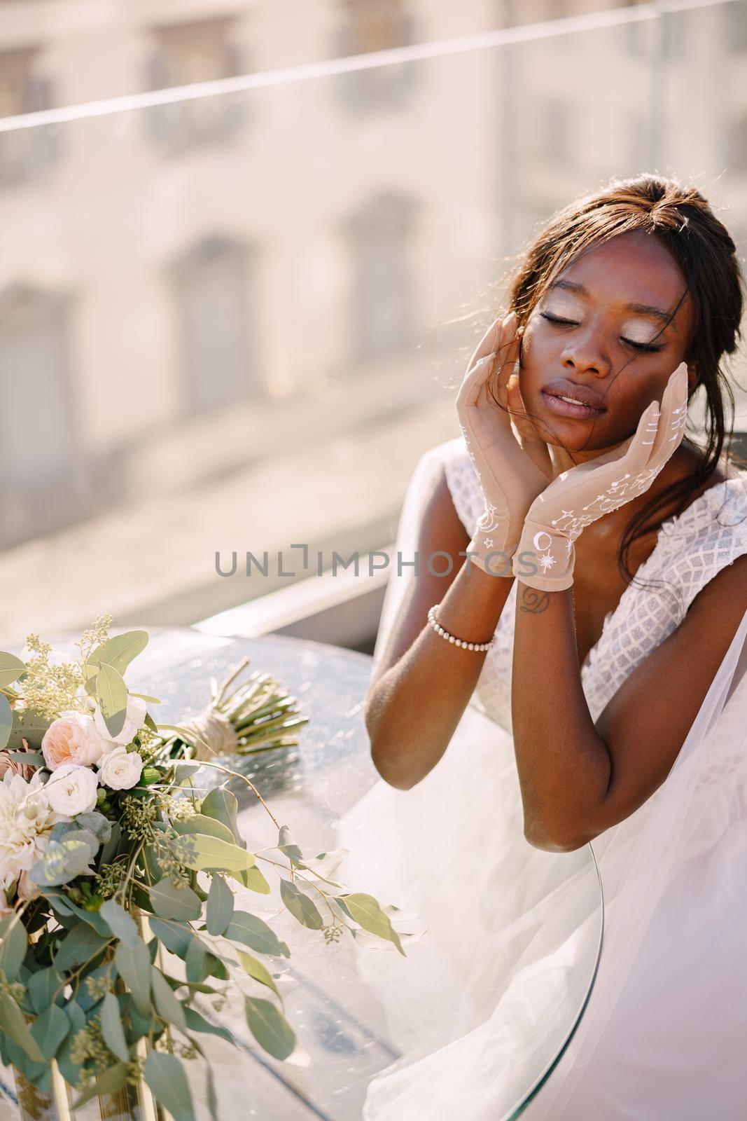 Destination fine-art wedding in Florence, Italy. African-American bride sits at the table, touches his face with her hands in gloves, a bouquet lies on the table.