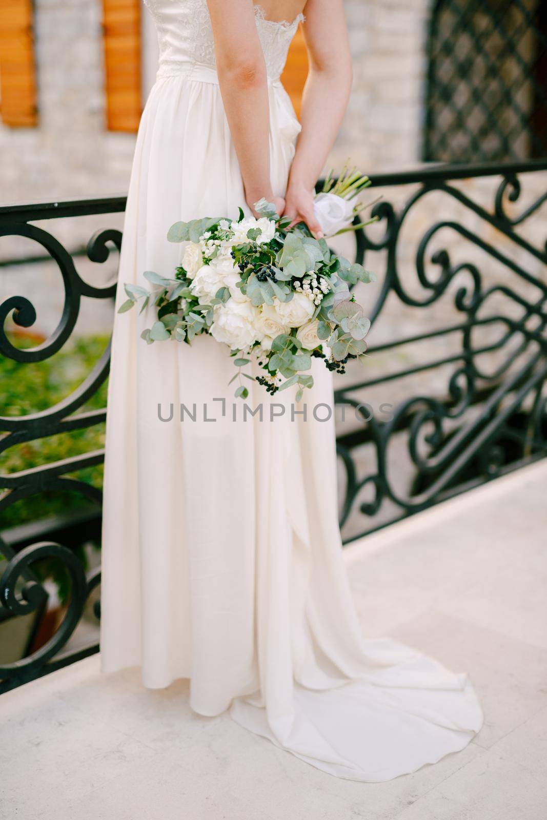 A graceful bride stands on a wrought-iron bridge in the old town of Budva and holds a bouquet behind her back, close-up by Nadtochiy