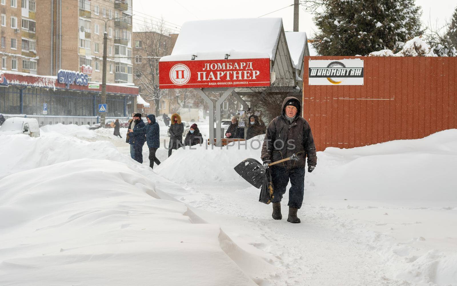 Lutsk, Ukraine - February 12,2020: City street after blizzard. An elderly man with snow shovel in winter. People walk in snow drifts on uncleaned street. Sidewalk covered in snow after storm.