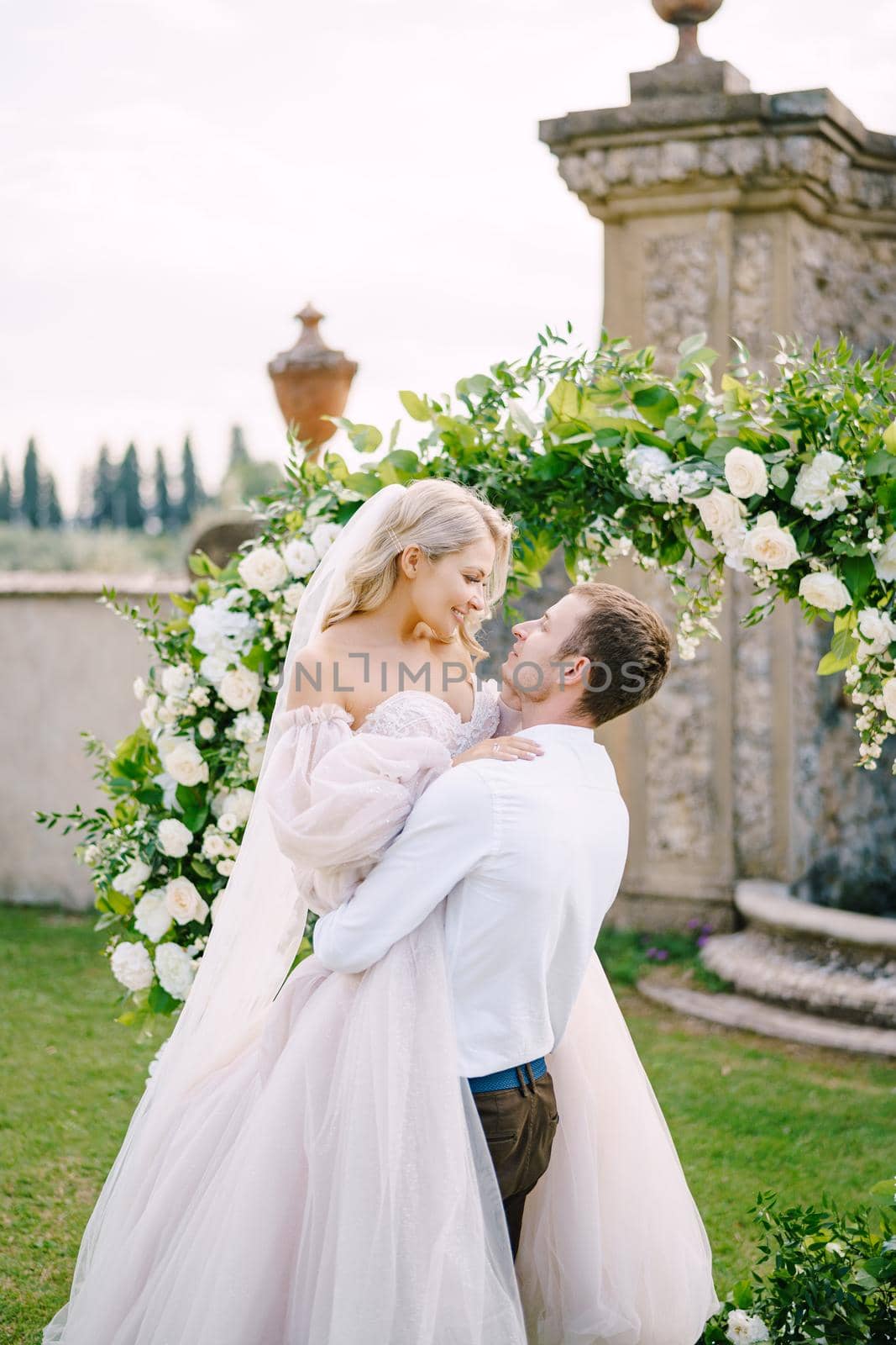 The groom is circling the bride in her arms. Round wedding arch decorated with white flowers and greenery in front of an ancient Italian architecture. Wedding at an old winery villa in Tuscany, Italy.