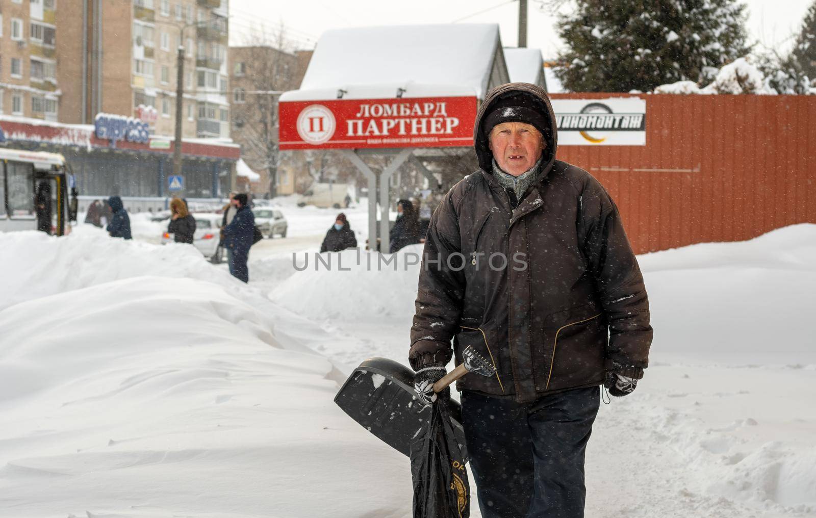 Lutsk, Ukraine - February 12,2020: City street after blizzard. An elderly man with snow shovel in winter. People walk in snow drifts on uncleaned street. Sidewalk covered in snow after storm.