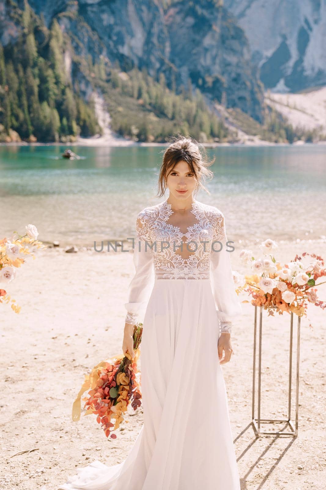 Beautiful bride in a white dress with sleeves and lace, with a yellow autumn bouquet on background of the arch for ceremony, at Lago di Braies in Italy. Destination wedding in Europe, on Braies lake. by Nadtochiy