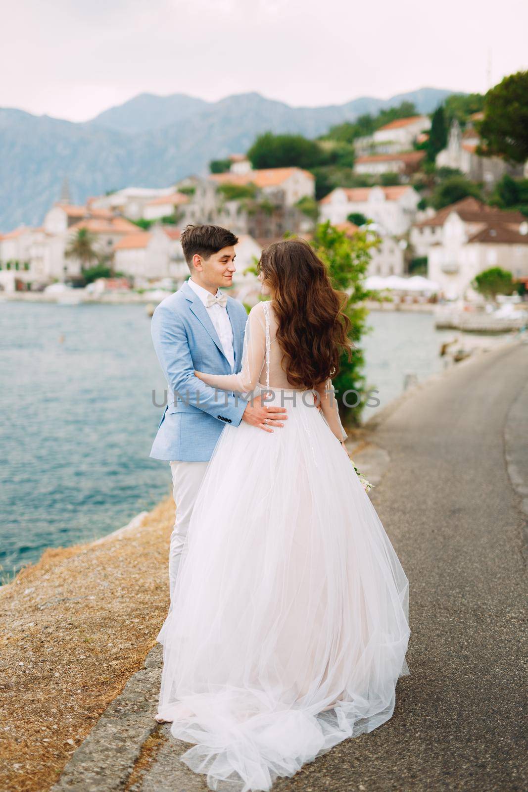 The bride and groom tenderly embrace on the seashore near the cozy old town of Perast in the Bay of Kotor . High quality photo