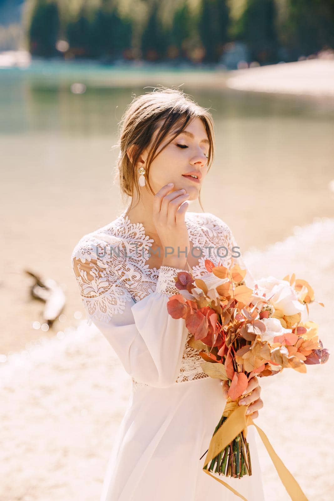 Beautiful bride in a white dress with sleeves and lace, with a yellow autumn bouquet of dried flowers and peony roses, on the Lago di Braies in Italy. Destination wedding in Europe, on Braies lake. by Nadtochiy