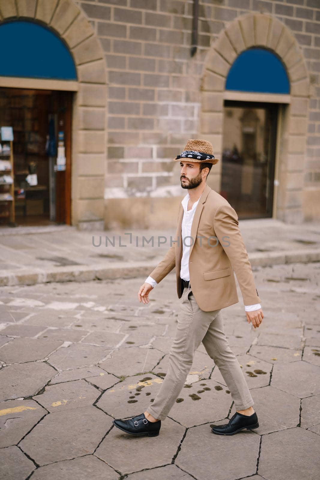 A young guy with a straw hat, a white unbuttoned shirt and a sand jacket, a small beard walks around the city of Florence. by Nadtochiy