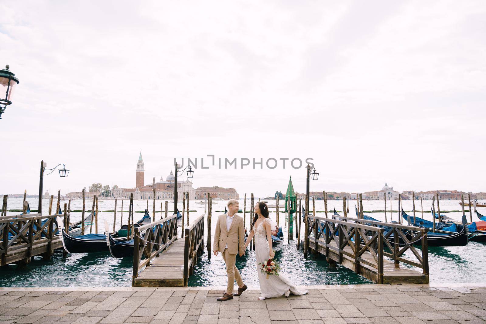 The bride and groom are walking along the gondola pier, holding hand in Venice, near Piazza San Marco, overlooking San Giorgio Maggiore and the sunset sky. The largest gondola pier in Venice, Italy. by Nadtochiy