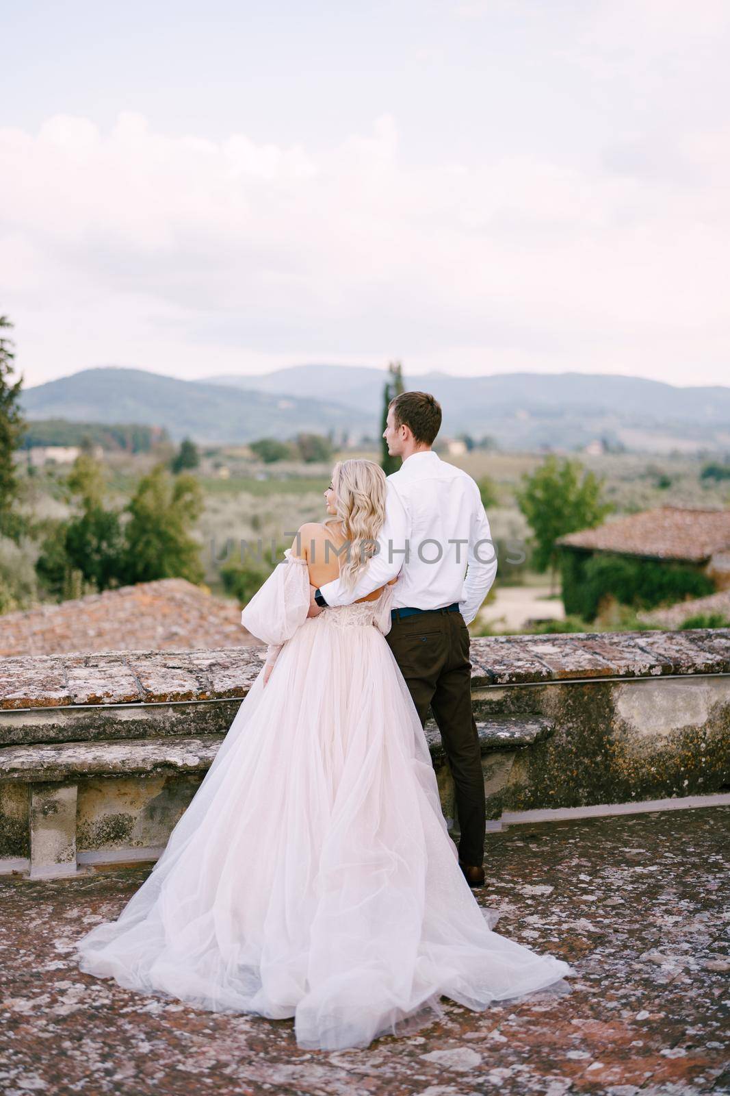 Wedding at an old winery villa in Tuscany, Italy. The wedding couple stands on the roof of an old winery, cuddling, standing with their backs in the frame.