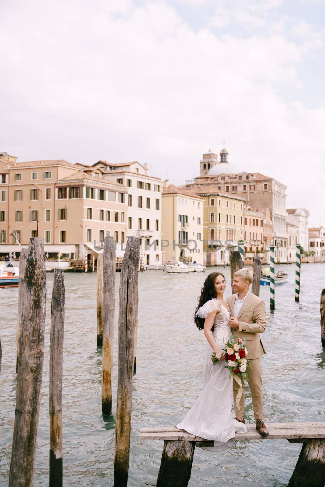 Italy wedding in Venice. The bride and groom are standing on a wooden pier for boats and gondolas, near the Striped green and white mooring poles, against backdrop of facades of Grand Canal buildings. by Nadtochiy
