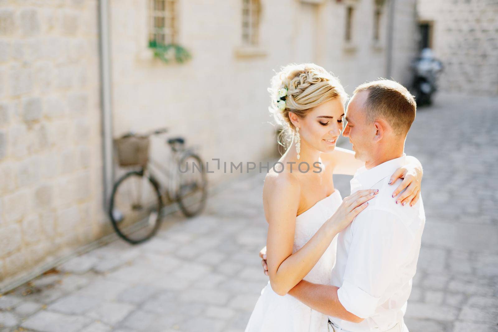 The bride and groom hug on the street of the old town of Perast next to a white building and a bicycle . High quality photo