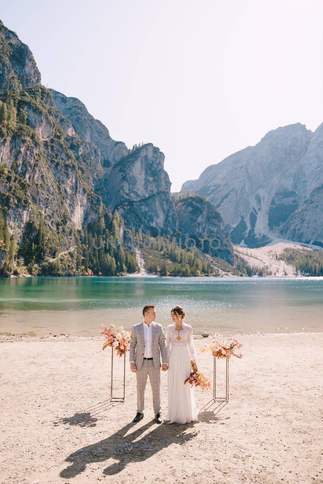 The bride and groom stand still for the ceremony, with an arch of autumn floral columns, against the backdrop of Lago di Braies in Italy. Destination wedding in Europe, at Braies lake.
