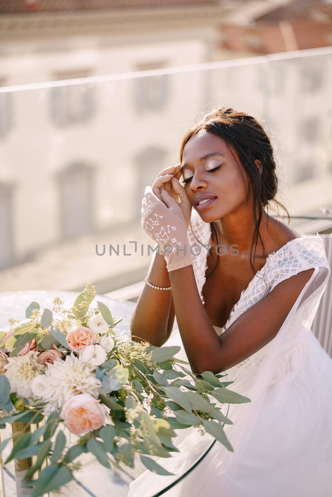 Destination fine-art wedding in Florence, Italy. African-American bride sits at the table, touches his face with her hands in gloves, a bouquet lies on the table.