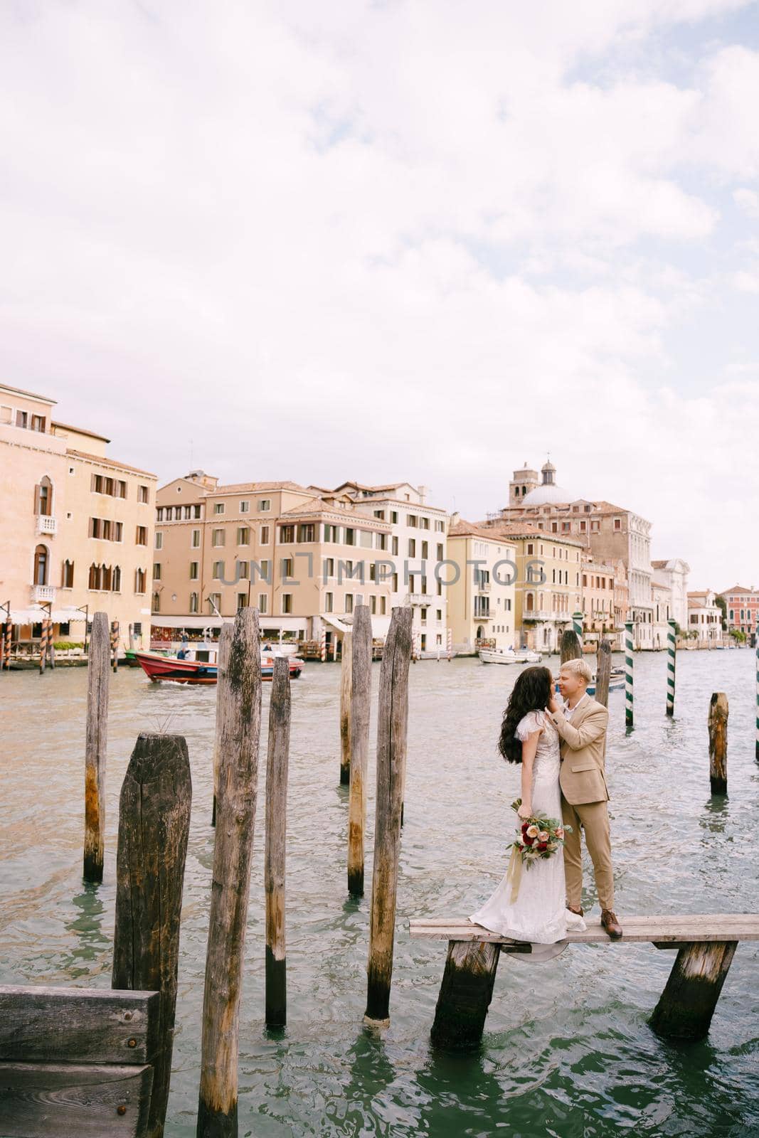 The bride and groom stand on a wooden dock for boats and gondolas, near striped green and white mooring poles, against the facades of the Grand Canal buildings.